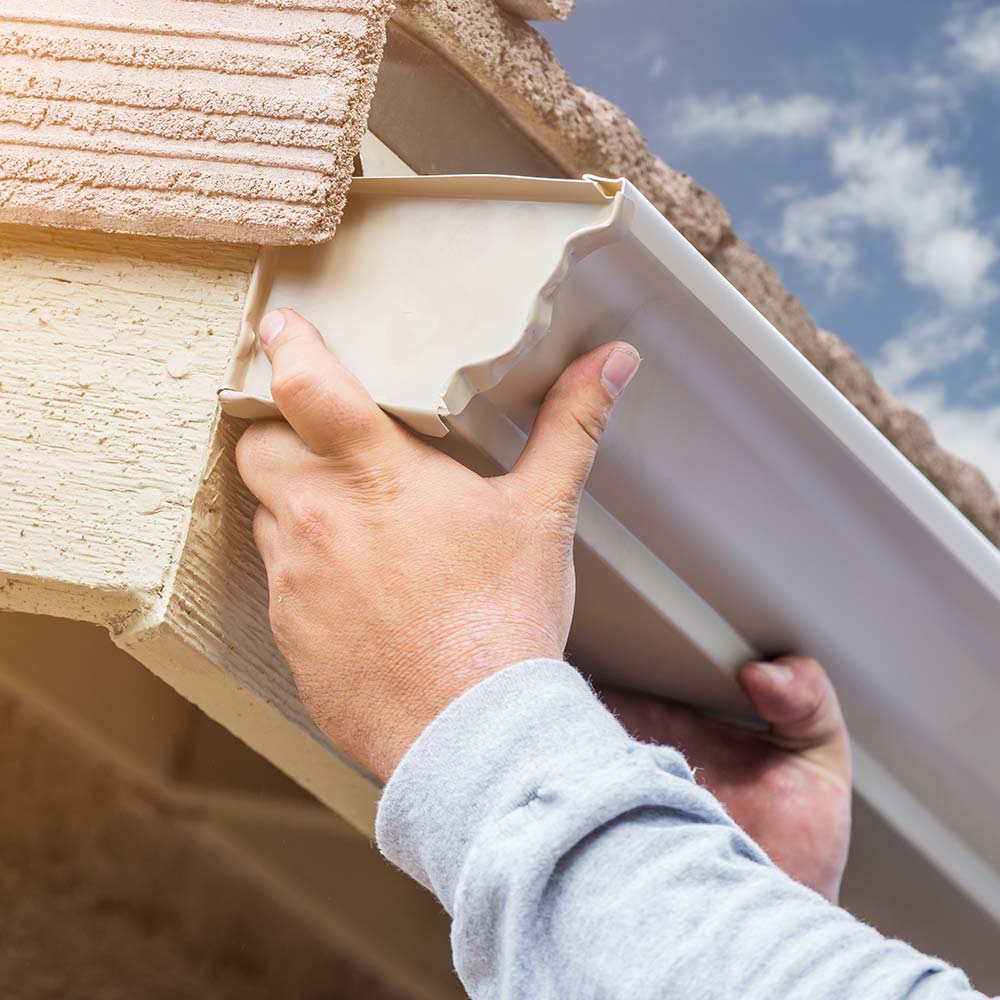 Hands in a gray sleeve attach a cream-colored gutter to a beige roof under a blue sky. The image conveys a sense of focus and craftsmanship.