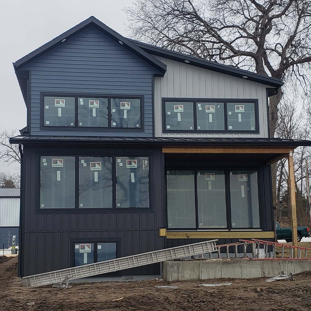 Two-story house under construction with dark blue and gray siding, large windows, and bare trees in the background. A ladder leans against the concrete foundation.