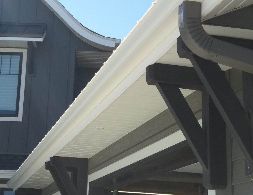 Close-up of a house's exterior showing white gutters and downspouts against dark gray siding. A clear blue sky is in the background.